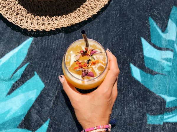 A hand holding a decorated drink with a straw, next to a straw hat on a tropical-themed background.