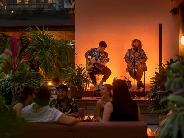 A cozy nighttime scene with live musicians performing outdoors; people are enjoying the ambiance surrounded by plants and soft lighting.