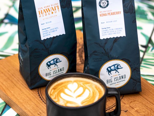 A latte with latte art in a black cup sits on a wooden board with two bags of coffee, featuring a leafy background.