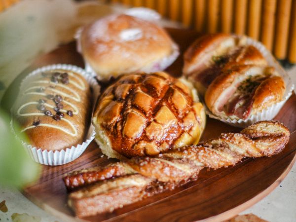 The image shows a wooden plate with a variety of baked goods, including bread rolls, pastries, and a breadstick twist, on a textured surface.