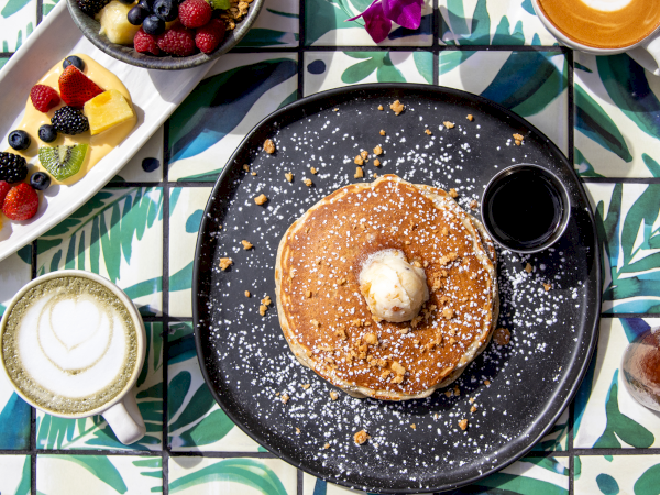 A plate with pancakes topped with butter and syrup, a side of fruits, and a cappuccino on a vibrant, patterned table.