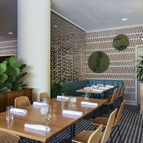 The image shows a stylish restaurant interior with wooden tables, chairs, plants, patterned walls, and napkins on the tables.