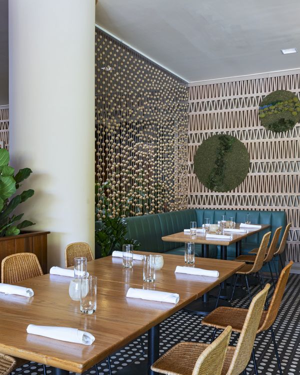 The image shows a stylish restaurant interior with wooden tables, chairs, plants, patterned walls, and napkins on the tables.