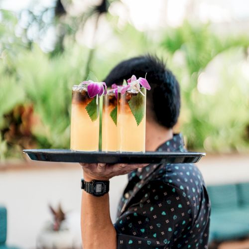 A server holds a tray with two tropical-looking drinks garnished with purple flowers, outdoors, person&rsquo;s face partly hidden.