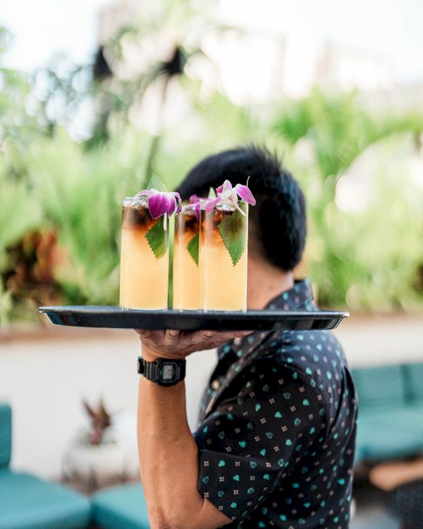 A server holds a tray with two tropical-looking drinks garnished with purple flowers, outdoors, person&rsquo;s face partly hidden.