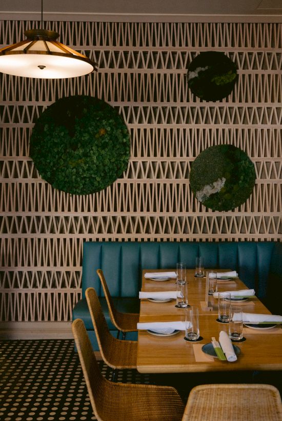 A modern restaurant interior with wooden tables, green seating, decorative wall patterns, circular plant accents, and overhead lighting.