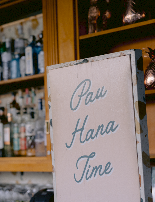 A bar scene with a sign saying "Pau Hana Time" and shelves stocked with various bottles and glassware in the background.