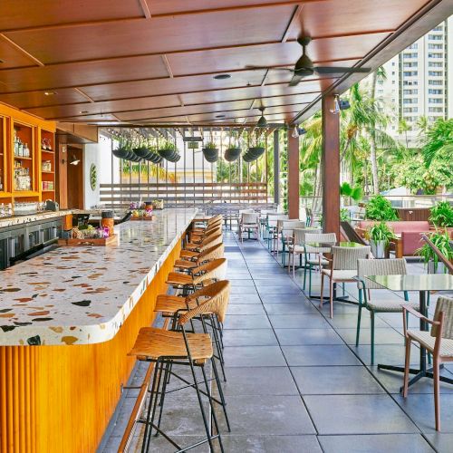 The image shows a modern outdoor bar with terrazzo countertops, wooden accents, high stools, tables, chairs, and a view of a cityscape in the background.