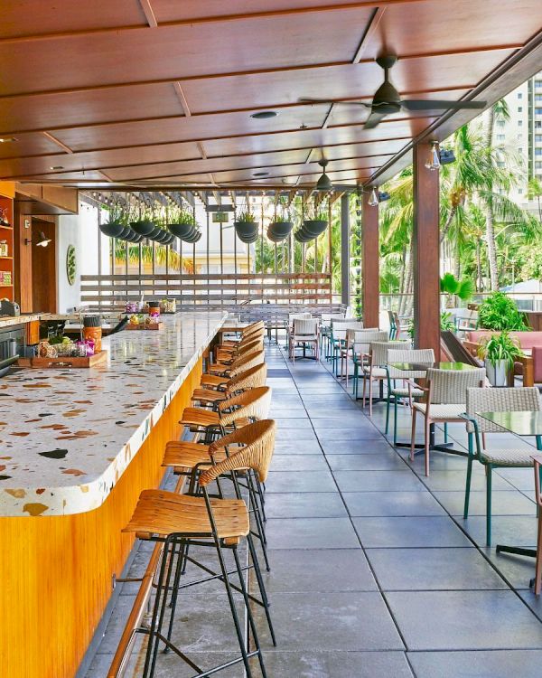 The image shows a modern outdoor bar with terrazzo countertops, wooden accents, high stools, tables, chairs, and a view of a cityscape in the background.