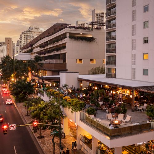 People dining on a terrace of a modern building beside a busy street at sunset, with city high-rises in the background.