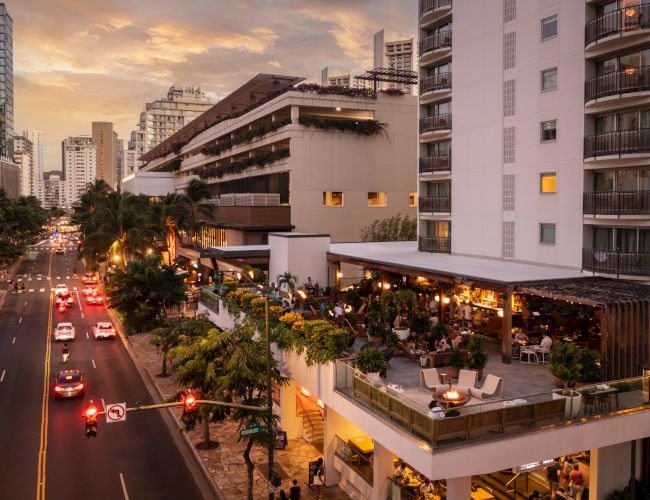 People dining on a terrace of a modern building beside a busy street at sunset, with city high-rises in the background.