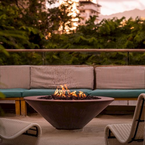 An outdoor seating area with a fire pit surrounded by cushioned benches and chairs, set against a backdrop of greenery and mountains.
