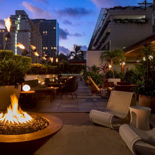 An outdoor lounge area with a fire pit, cozy seating, lush plants, and surrounding buildings against a dusk sky.